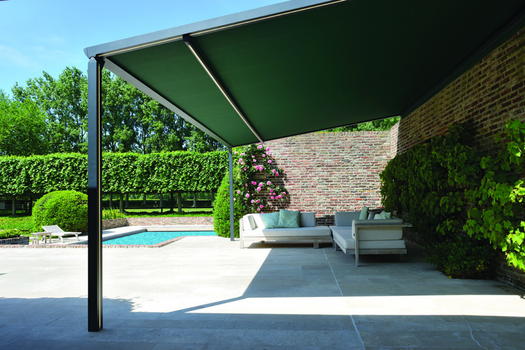 Modern outdoor patio with green awning, cushioned seating, brick wall with climbing plants, and poolside view.