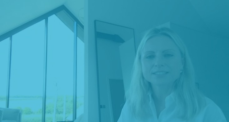 A woman stands in a serene, modern master bedroom with large architecturally shaped glazing. The room is softly tinted in WindowTreat Blue, showcasing the Trapezia electric lifting roller blind discreetly covering the expansive 5.5m x 4.5m window.