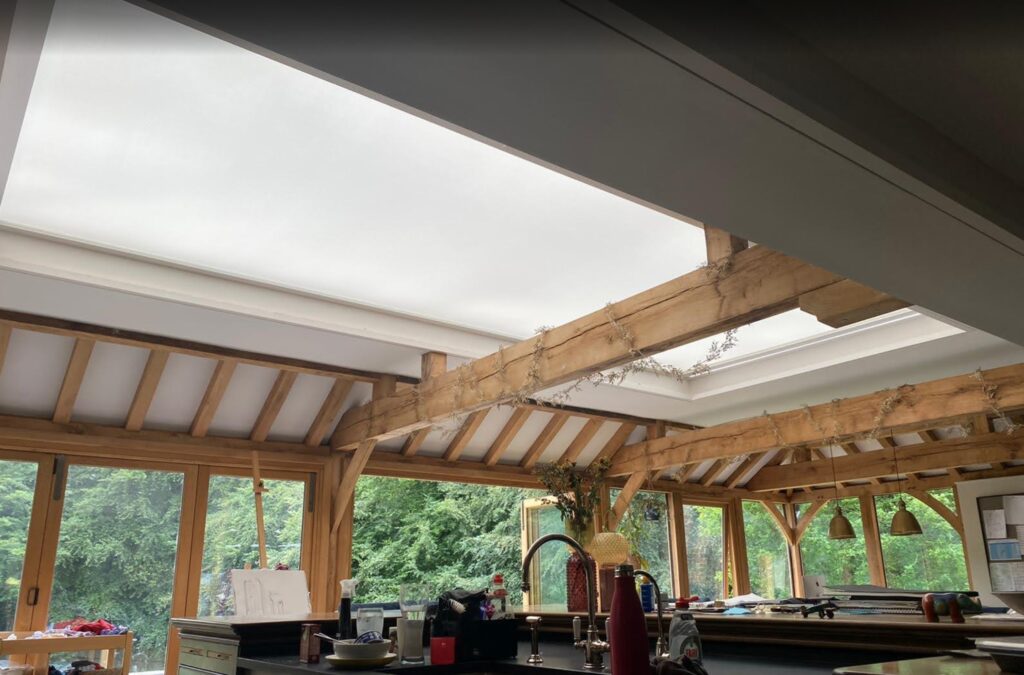 Large Hercules roof blind with white fabric, filtering sunlight through a skylight in a forest-facing kitchen.