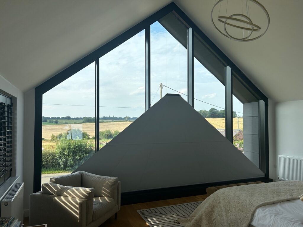 Architectural master bedroom with large triangular glazing overlooking fields, featuring minimalist furnishings and natural light.