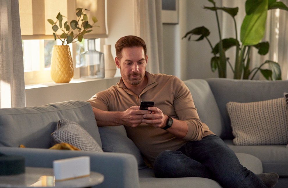 Man sitting on a light blue sectional sofa in a sunlit living room, using his smartphone to control window treatments via the Somfy TaHoma app.