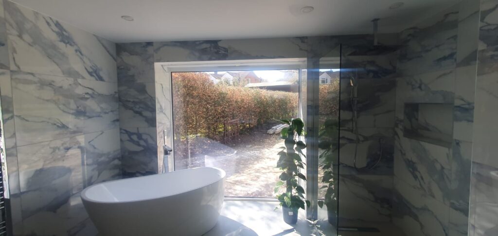 A modern bathroom with a large glass door opening to a garden. An electric privacy blind is concealed using Blindspace® at the top of the window surround, preserving the uninterrupted view and glazing.
