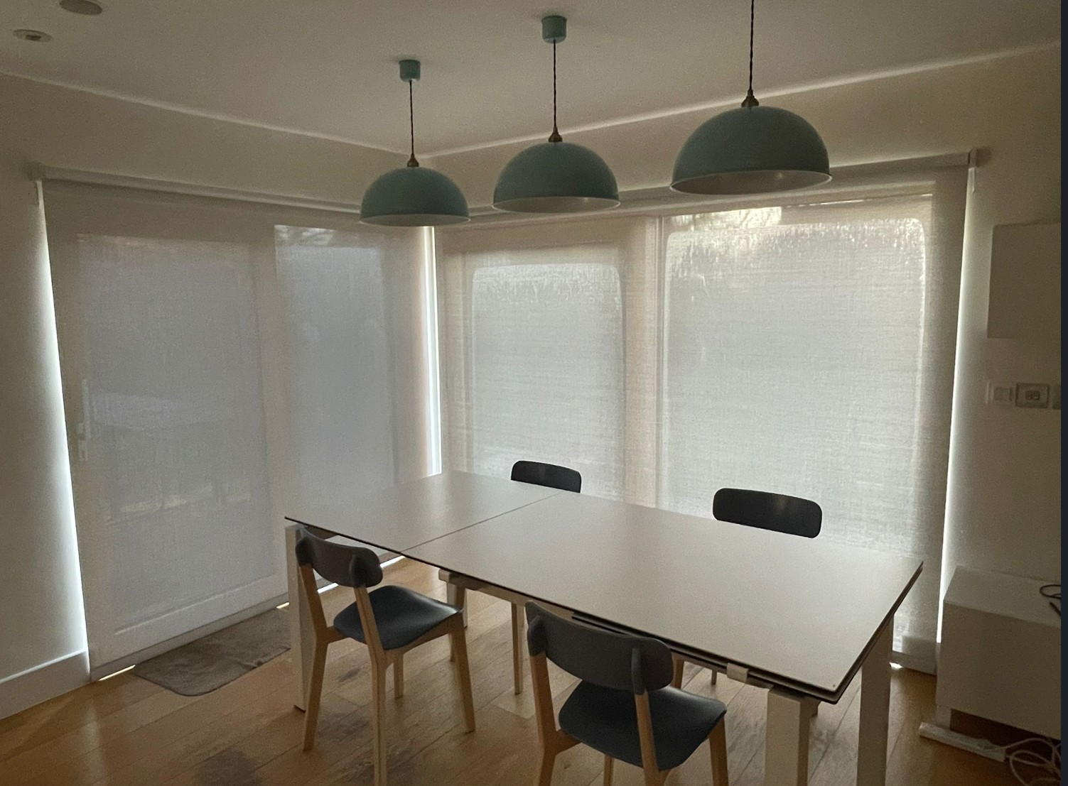 Modern dining room with large windows fitted with white roller blinds, diffusing natural light across a minimalist table and chairs.