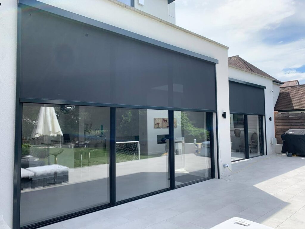 Exterior view of a modern home with large glass doors covered by dark roller blinds, fitted with zipped side channels for sun protection and privacy.