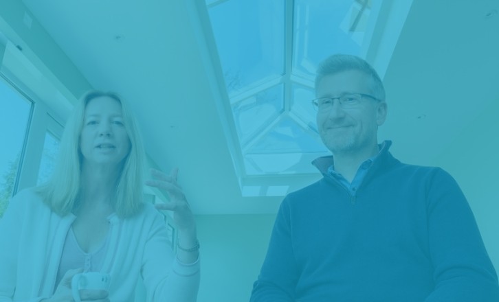 Two homeowners sit beneath a large roof lantern in their new extension, talking together in a bright modern room with concealed blinds.